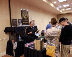 #image_title Man in a black shirt selling books at a vendor table in a conference room with posters behind him.