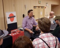 #image_title Magician in a purple shirt performing a card trick for a small audience at a booth with Paul Richards Magic sign on display table.