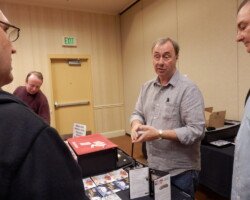 #image_title Man in gray button-down shirt demonstrating or handling cards at a vendor table in a conference room, with people watching nearby and an exit sign in the background.