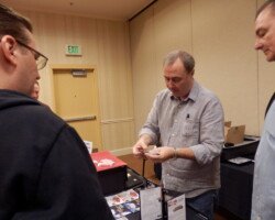 #image_title Man in a gray shirt shows cards at a vendor table during an indoor conference or trade show.